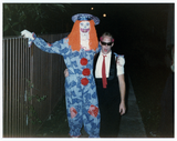 Man dressed as clown with man in sunglasses by fence at a Halloween party